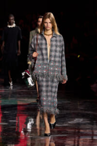 Model walks the runway in a gray plaid fringed suit with a beige top, gold chain necklace, and black patent heels.