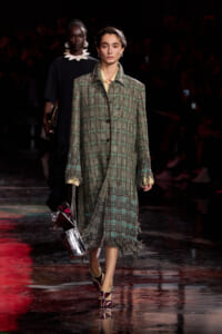 Model walks the runway in a green plaid overcoat with fringe, carrying a metallic handbag and wearing strappy green heels.
