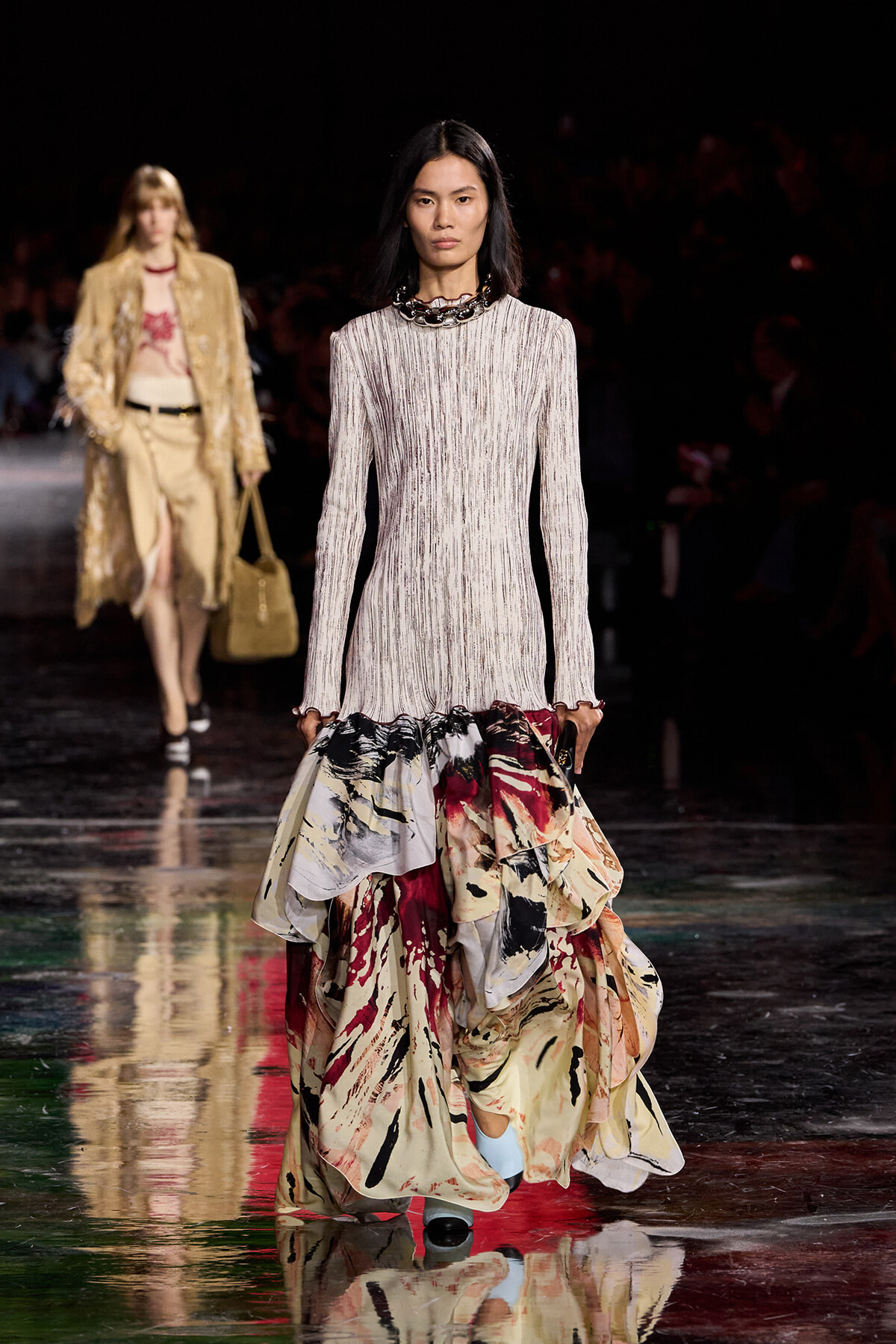 Model walks a fashion runway in a long-sleeve, light-striped top and a dramatic, multi-colored, ruffled skirt with an abstract print.