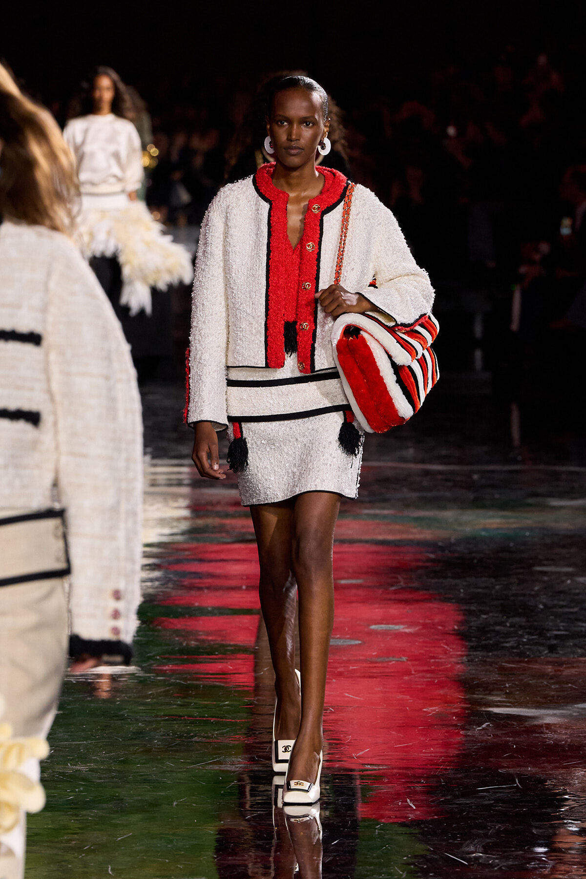 Model walking a runway in a white textured suit with red trim, carrying a red-and-white striped bag.