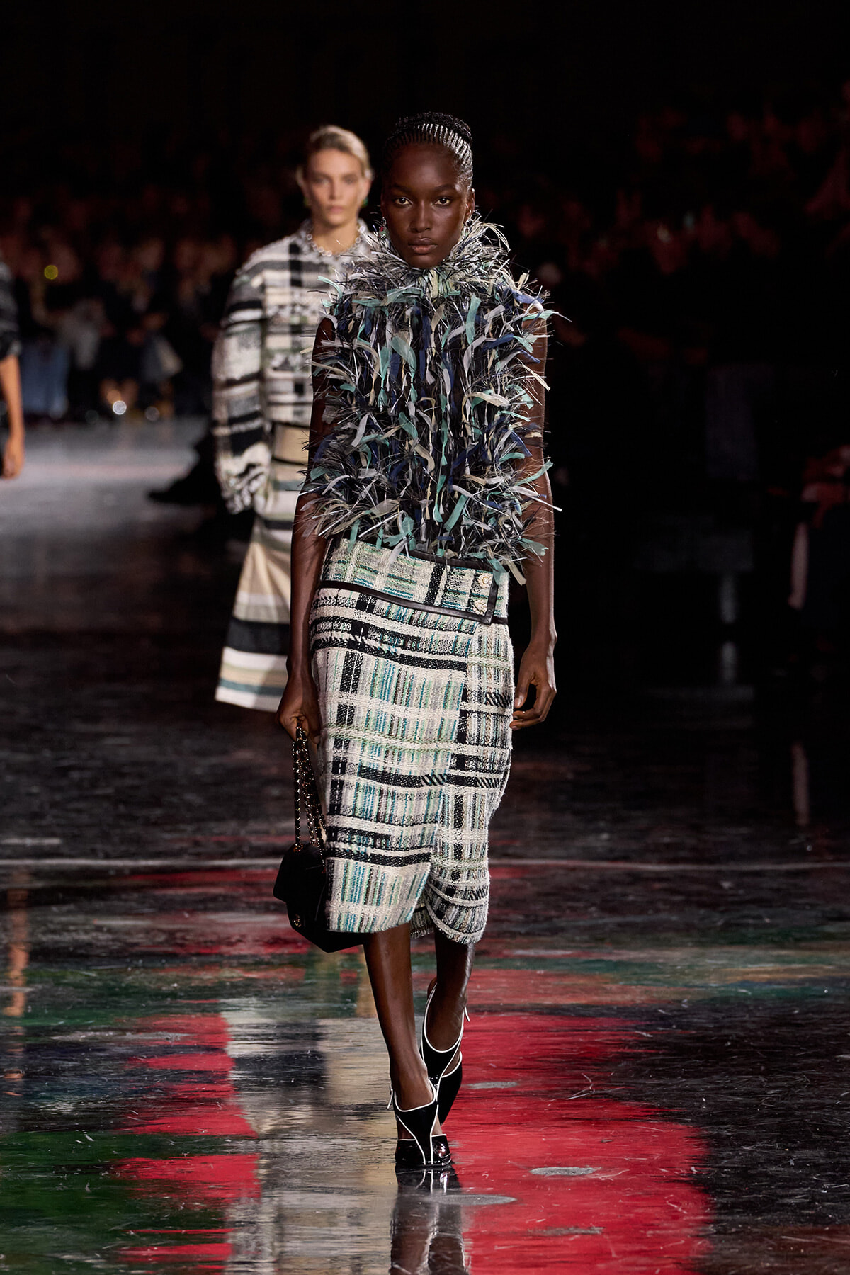 Model walks the runway in a sleeveless, plaid tweed dress with a voluminous feathered collar in teal and silver; accessorized with a small black handbag and black strappy heels.