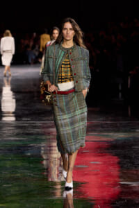 Model walks runway wearing a green plaid tweed skirt suit with red buttons and a mustard plaid top, white heels, and a metallic handbag.