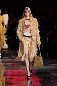 Runway model in a fringed beige coat over a sheer red floral top, beige skirt with a slit, black belt, and tan handbag.