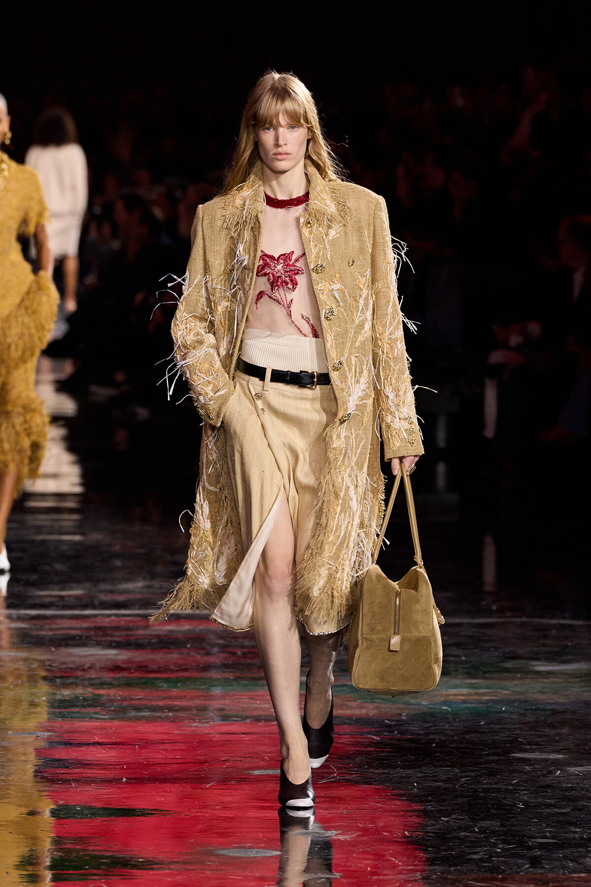 Runway model in a fringed beige coat over a sheer red floral top, beige skirt with a slit, black belt, and tan handbag.