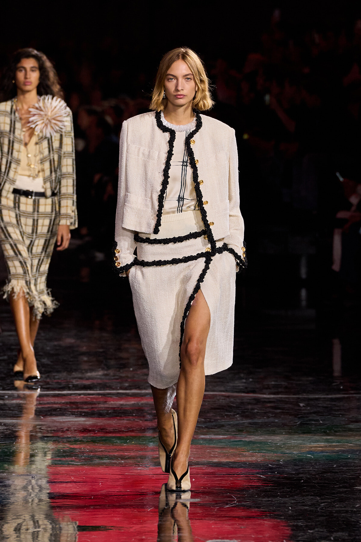 Model walking a runway in a cream tweed suit with black trim and gold buttons; slit skirt reveals her leg, black heels.