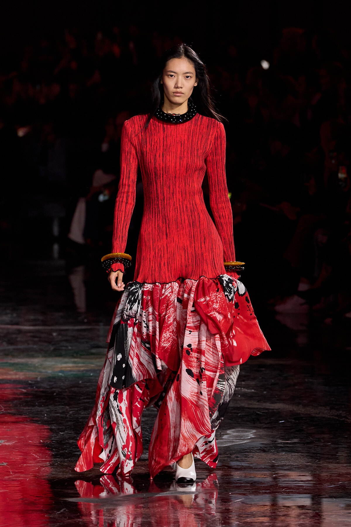 Female model walks a runway in a red knit dress with a black beaded collar and a voluminous red‑white‑black printed skirt, wearing stacked bracelets.