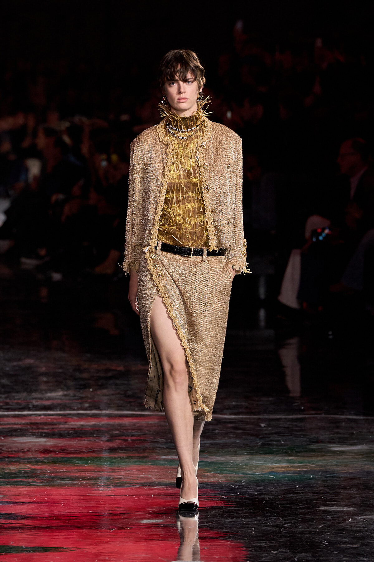 Model walking a fashion show in a gold tweed suit with a high-slit skirt and matching jacket, accessorized with layered gold necklaces.
