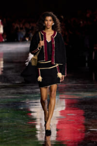 Model walking a fashion runway in a black boucle suit with burgundy trim, tassels, and gold buttons, carrying a tan shoulder bag.