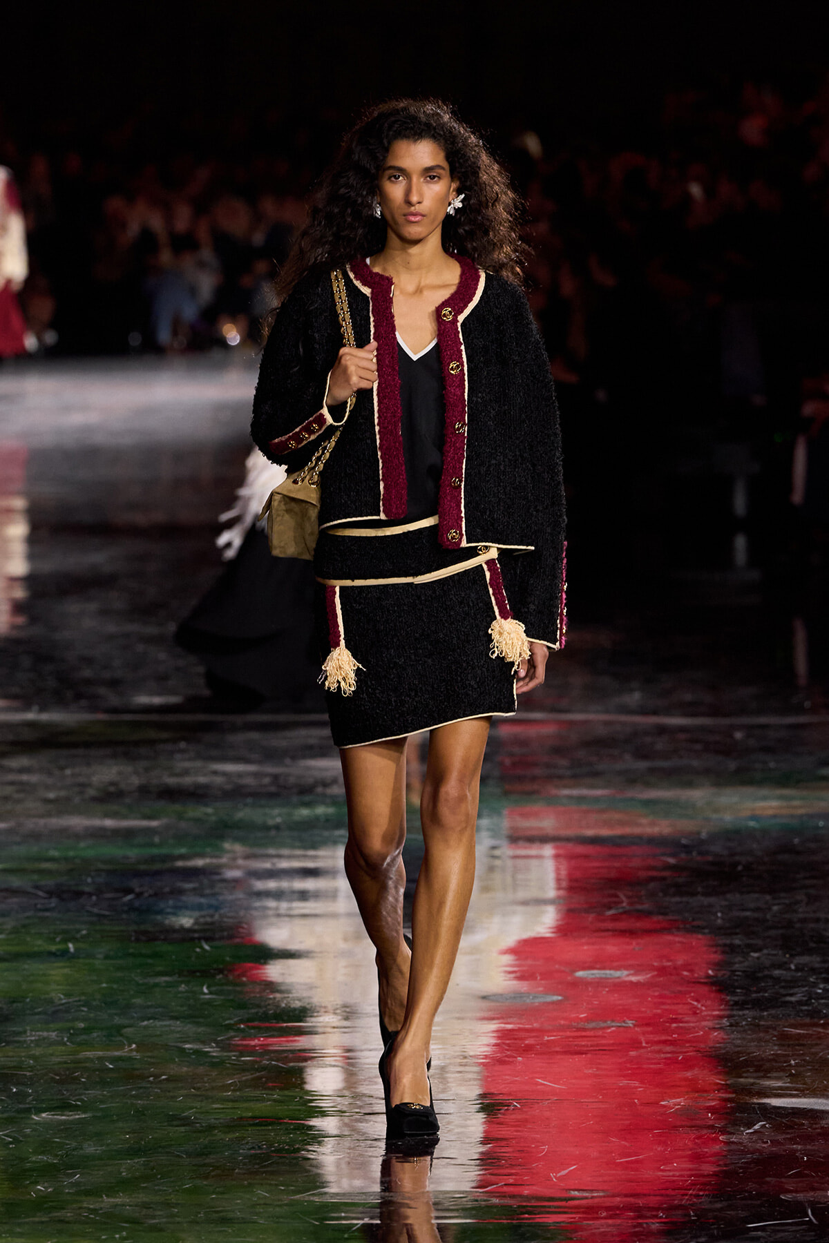 Model walking a fashion runway in a black boucle suit with burgundy trim, tassels, and gold buttons, carrying a tan shoulder bag.