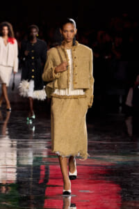 Model walks the runway in a gold tweed two-piece suit with a white blouse and gold trim details, carrying a chain-strap bag.