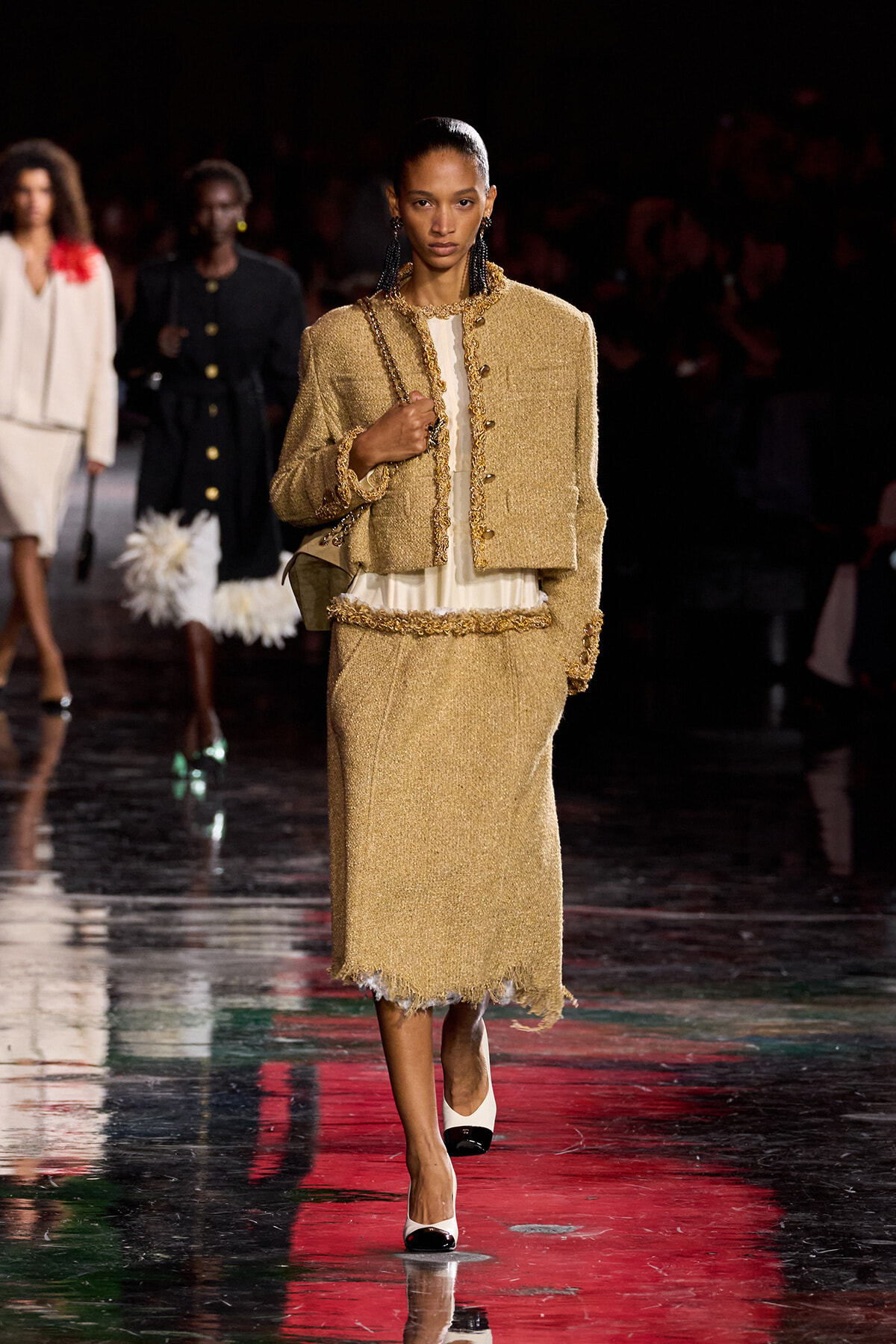 Model walks the runway in a gold tweed two-piece suit with a white blouse and gold trim details, carrying a chain-strap bag.