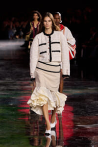 Model walks the runway wearing an ivory boucle jacket with black trim and matching layered cream skirt with a voluminous ruffled hem.