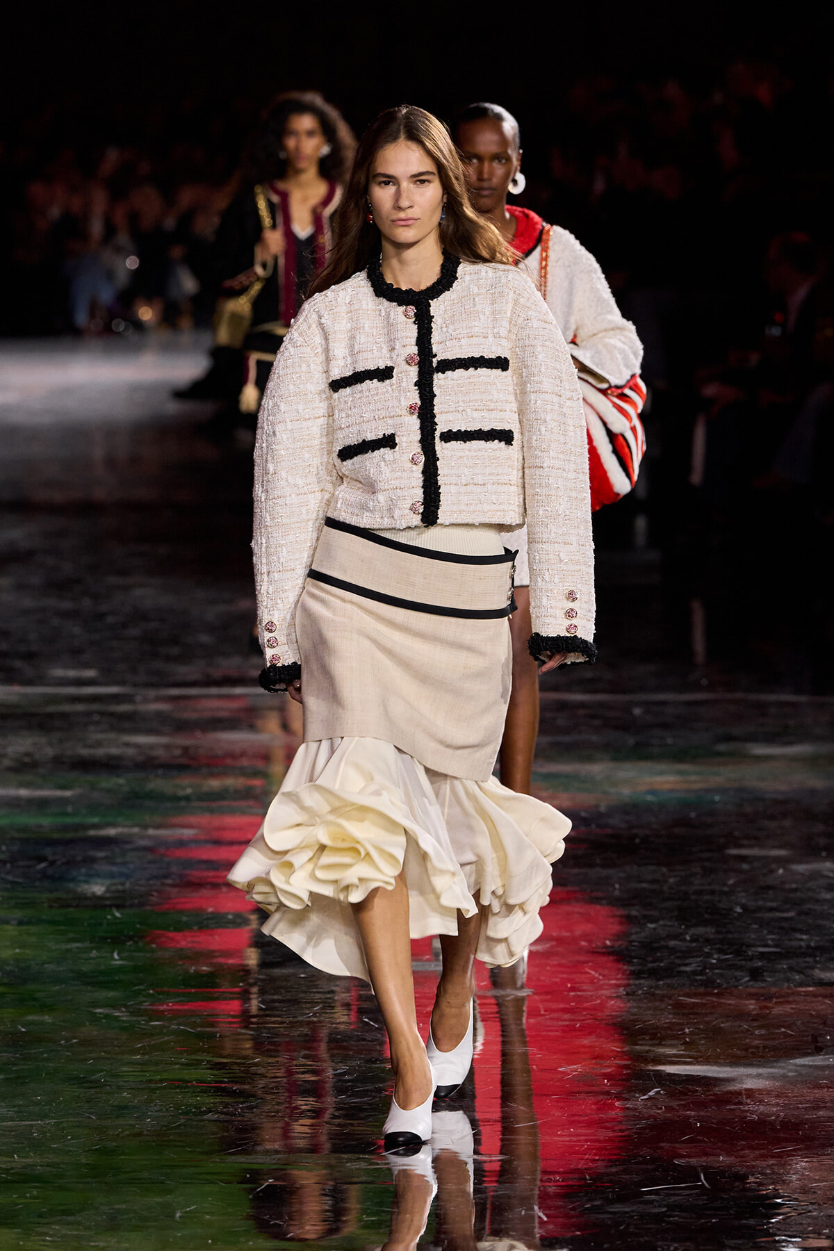 Model walks the runway wearing an ivory boucle jacket with black trim and matching layered cream skirt with a voluminous ruffled hem.