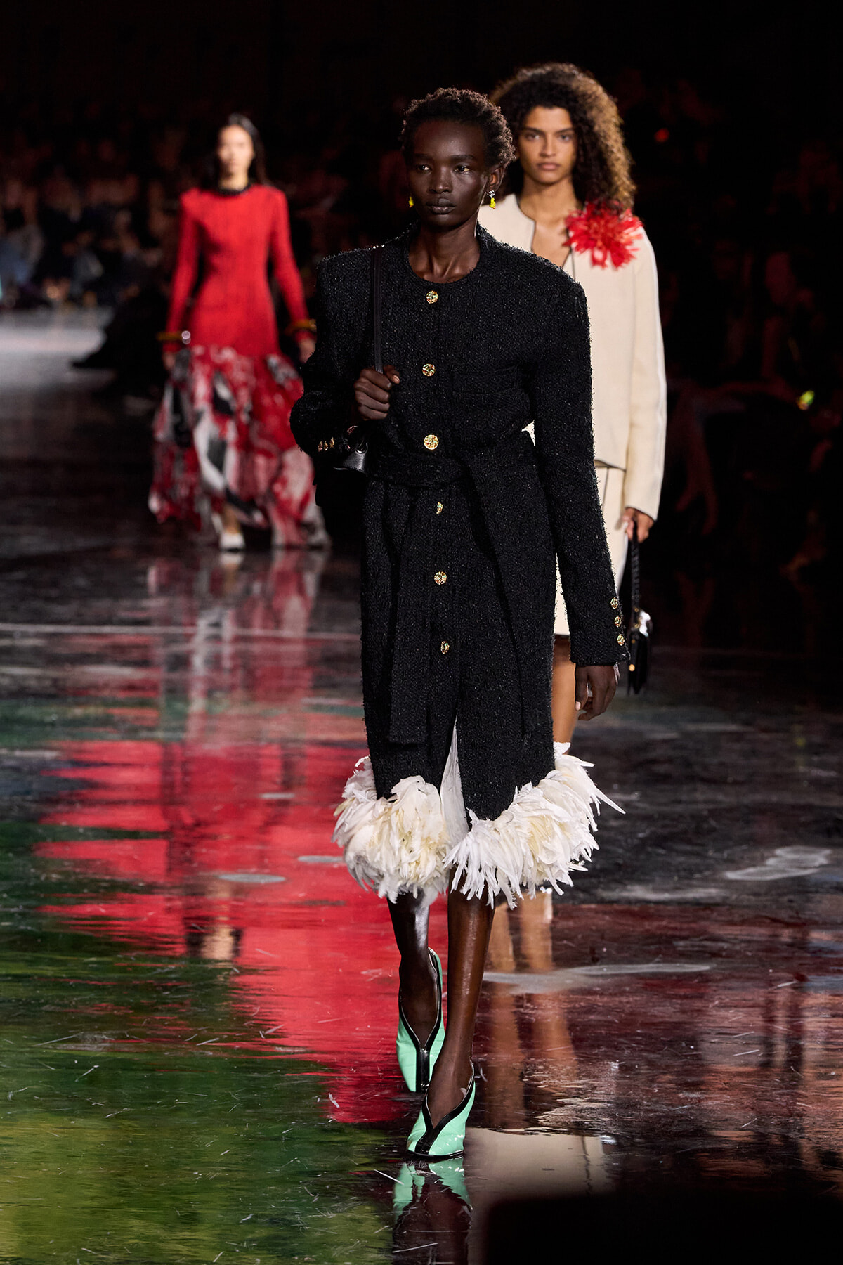 Model walking the runway in a black textured coat with gold buttons and white feathered hem, mint green pumps.