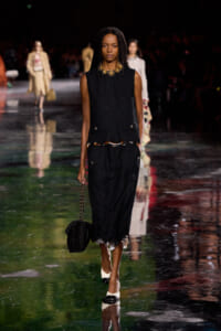 Model walks a fashion runway in a black textured sleeveless top and skirt with fringe, wearing a gold statement necklace and carrying a black handbag with a chain.