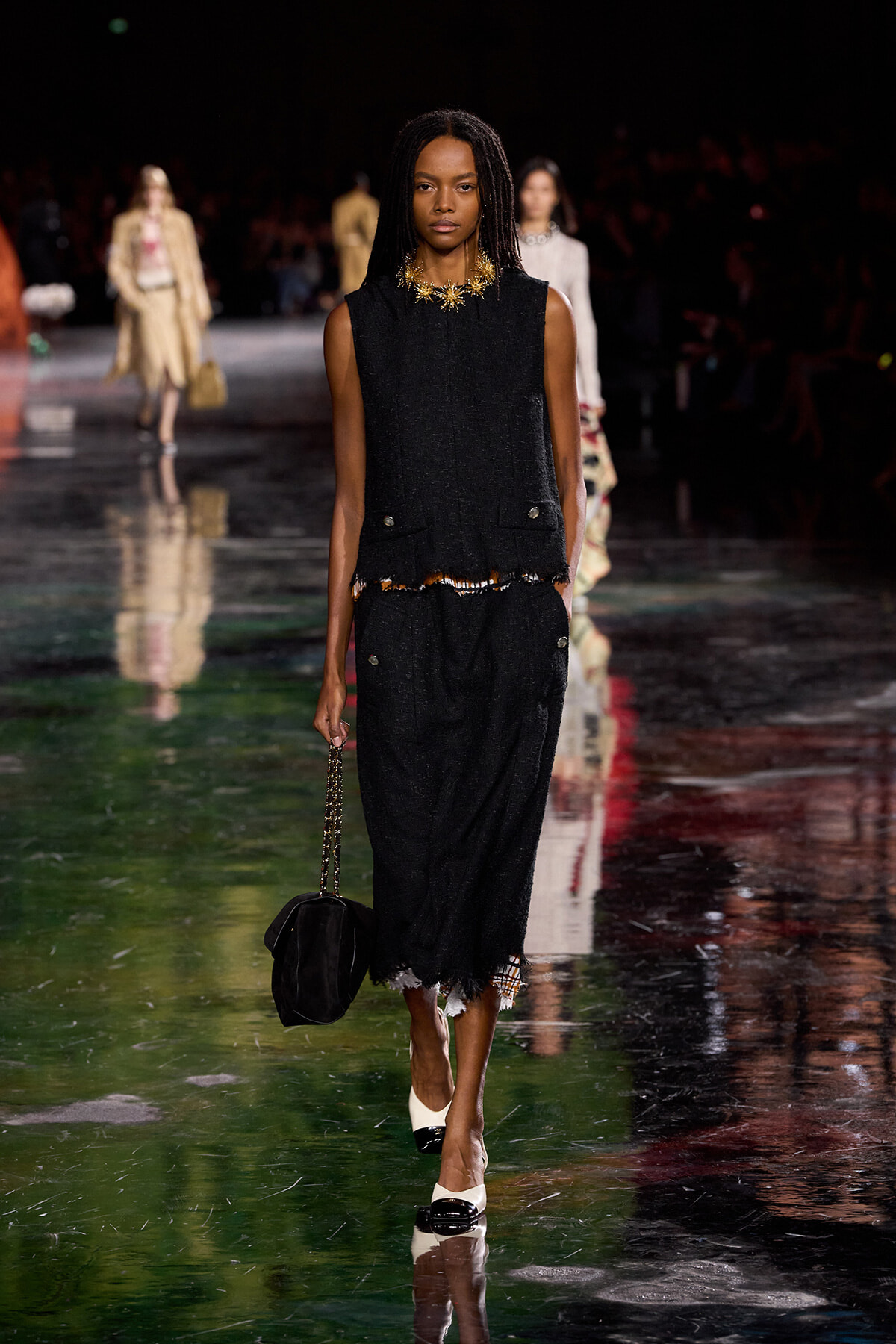 Model walks a fashion runway in a black textured sleeveless top and skirt with fringe, wearing a gold statement necklace and carrying a black handbag with a chain.