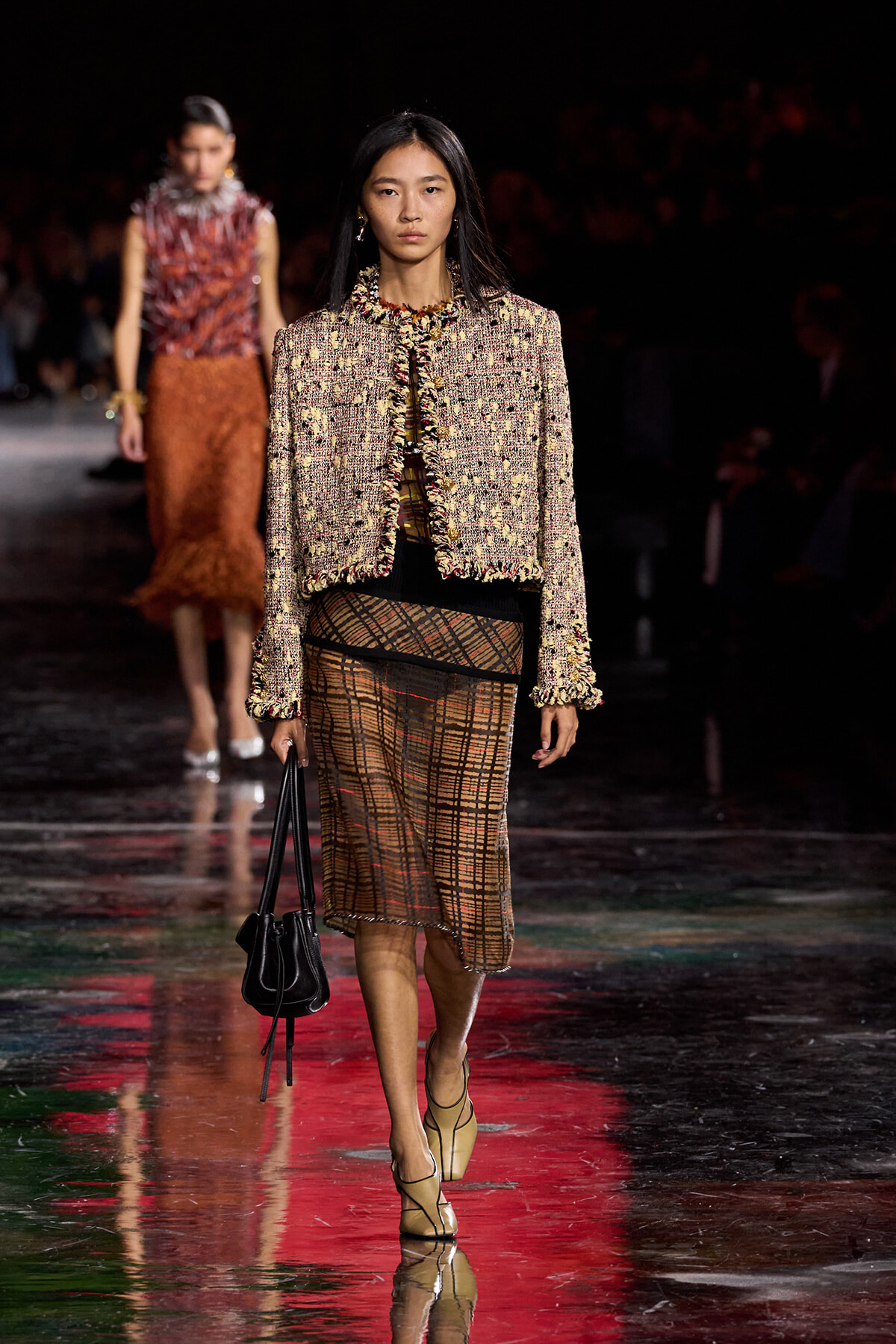 Model walking a fashion runway in a textured tweed jacket with fringe trim and a sheer plaid skirt, carrying a black handbag.