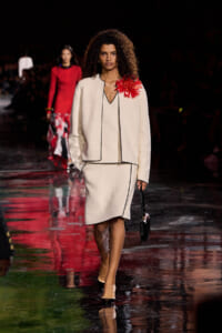 Model walks a cream suit with black piping on a glossy runway; red flower adorns her shoulder, holds a small black handbag.
