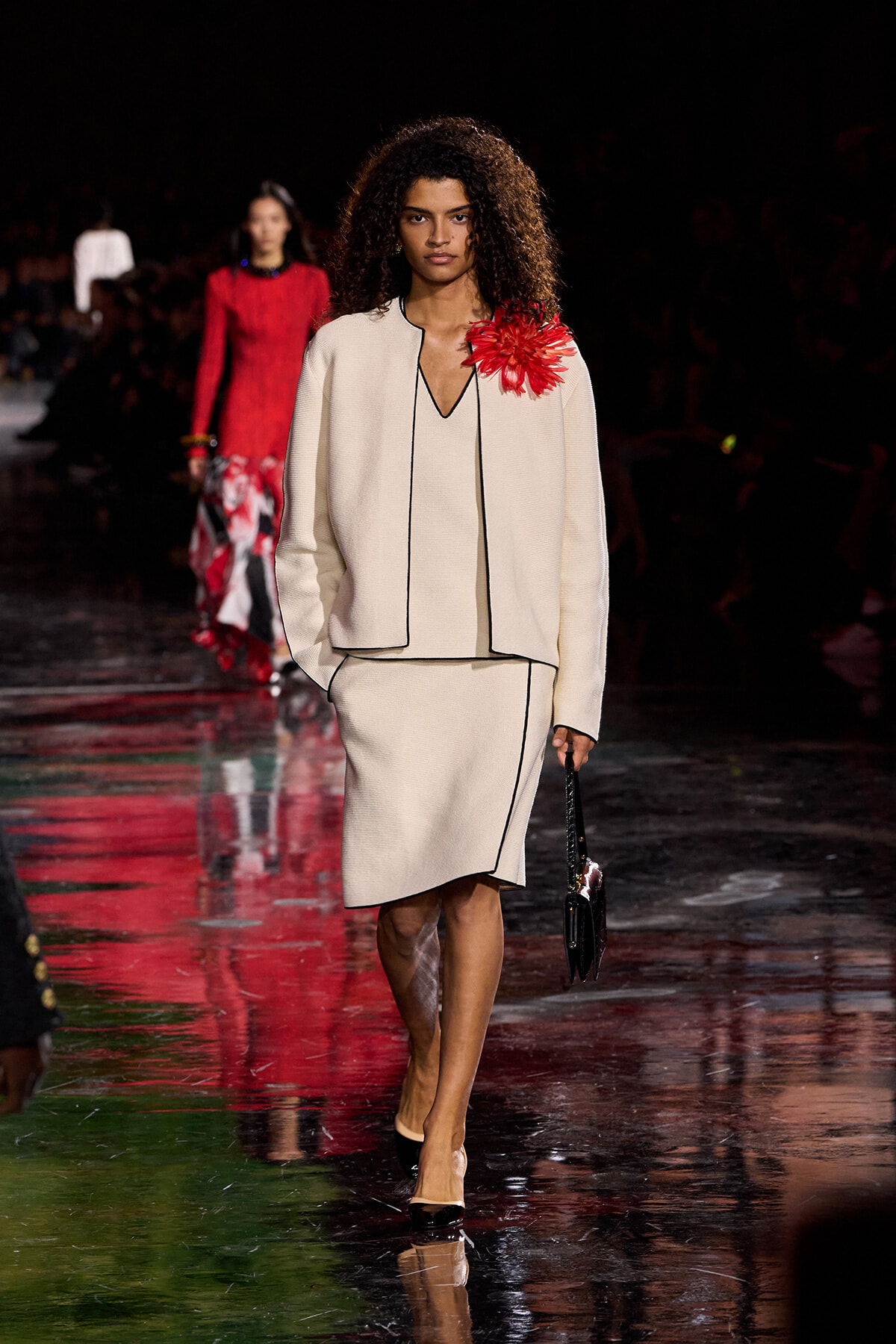Model walks a cream suit with black piping on a glossy runway; red flower adorns her shoulder, holds a small black handbag.