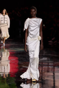 Model walks a white satin one-shoulder gown with draped sleeve and a side flower detail on a glossy runway.