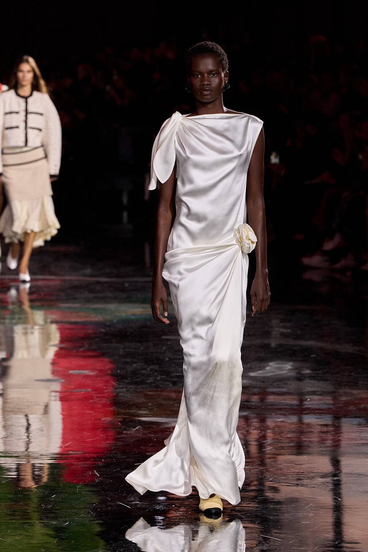 Model walks a white satin one-shoulder gown with draped sleeve and a side flower detail on a glossy runway.
