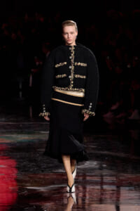 Model walking a runway in a black boucle jacket with gold braided trim and matching cuffs, paired with a layered black skirt and white-black heels.