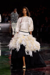 Model on a runway wearing an ivory satin blouse and a voluminous feathered white skirt over a black base, with cream heels.