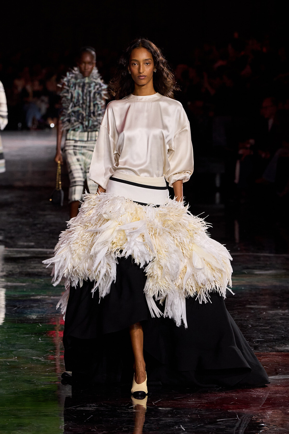 Model on a runway wearing an ivory satin blouse and a voluminous feathered white skirt over a black base, with cream heels.