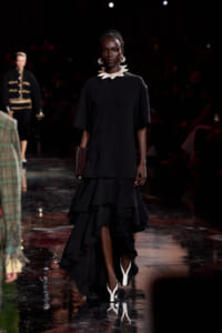 Dark-skinned model walking a runway in a black knee-length dress with a high-low ruffled skirt, pairing a statement white necklace and earrings with a burgundy clutch and white-heeled shoes.