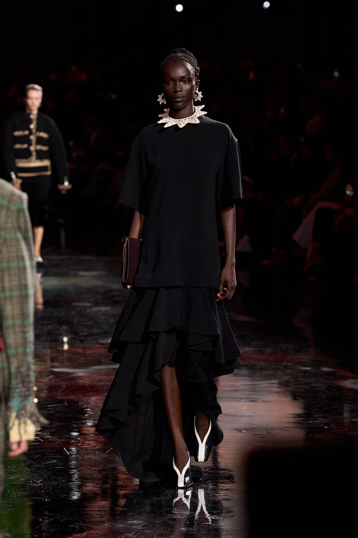 Dark-skinned model walking a runway in a black knee-length dress with a high-low ruffled skirt, pairing a statement white necklace and earrings with a burgundy clutch and white-heeled shoes.