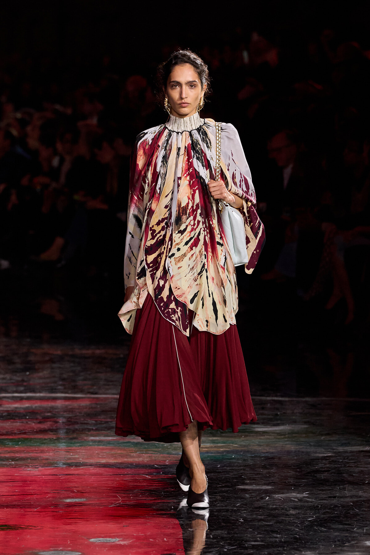 Model walking a fashion runway in a flowing multicolored abstract blouse with a high ruffled neck and burgundy pleated skirt, carrying a light blue bag, black heels.