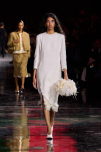 Model walks the runway in a white textured midi dress, carrying a fluffy white feathered clutch.