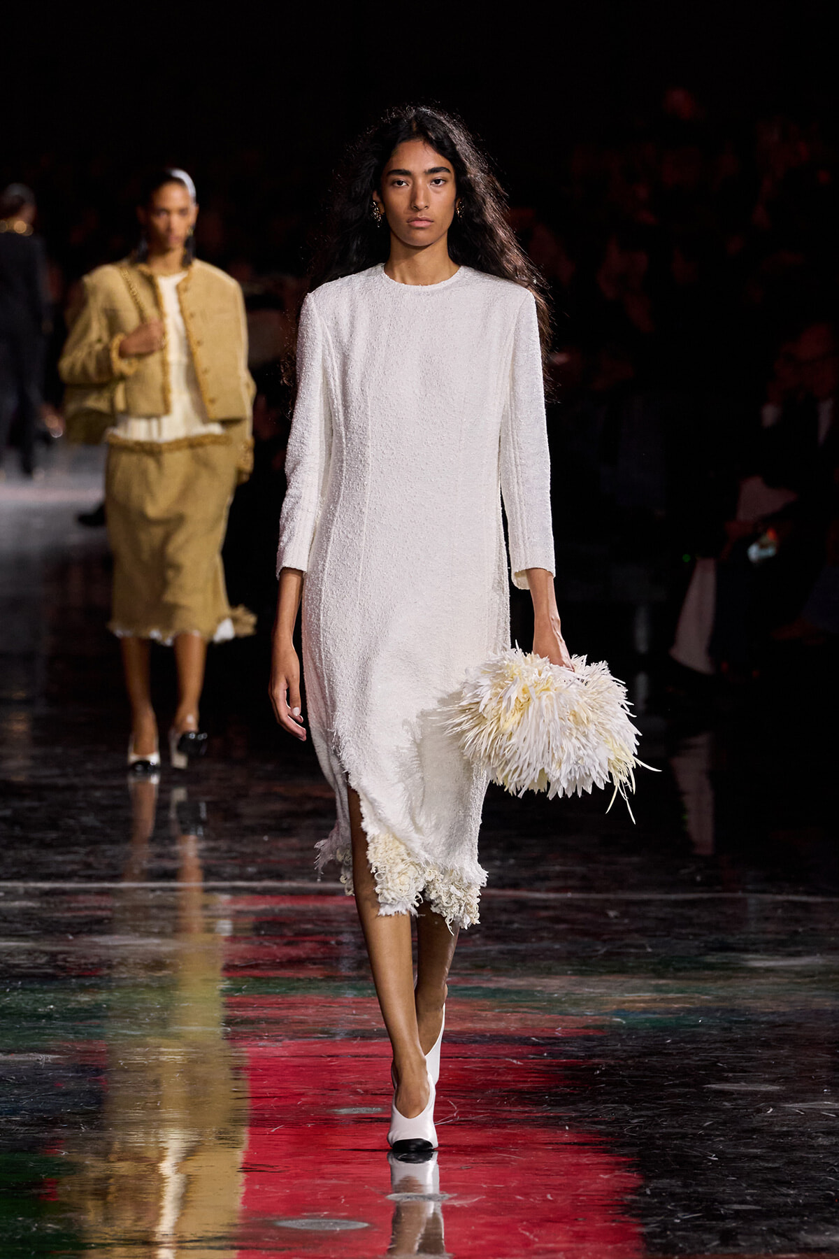 Model walks the runway in a white textured midi dress, carrying a fluffy white feathered clutch.