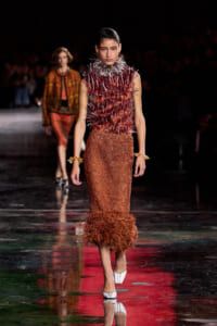 Model on a runway wearing a burgundy spiked top with a textured orange midi skirt and white heels, under dark venue lighting.
