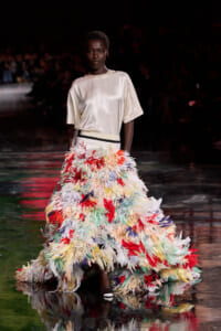 Model walks the runway in a cream satin top paired with a dramatic multicolor feather skirt that sweeps the floor.