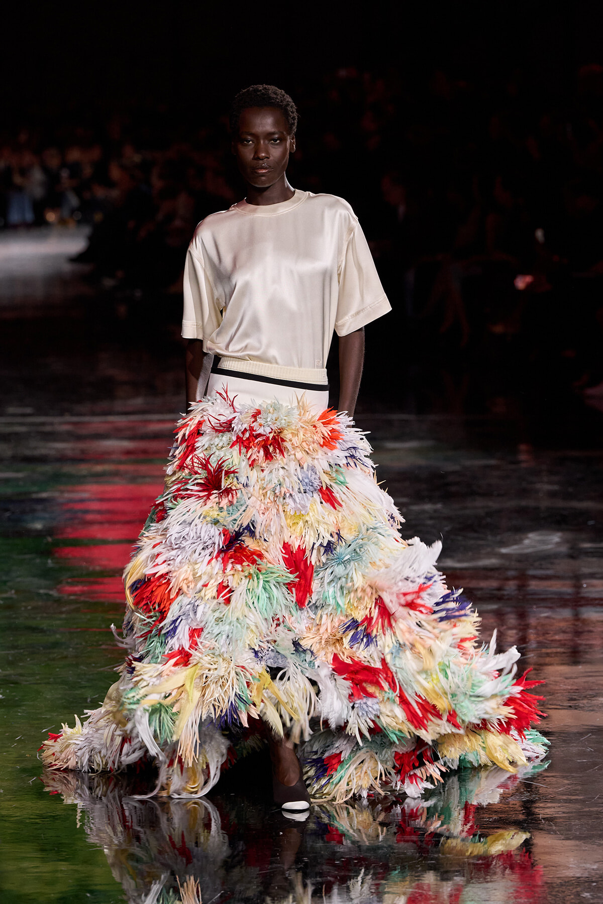 Model walks the runway in a cream satin top paired with a dramatic multicolor feather skirt that sweeps the floor.