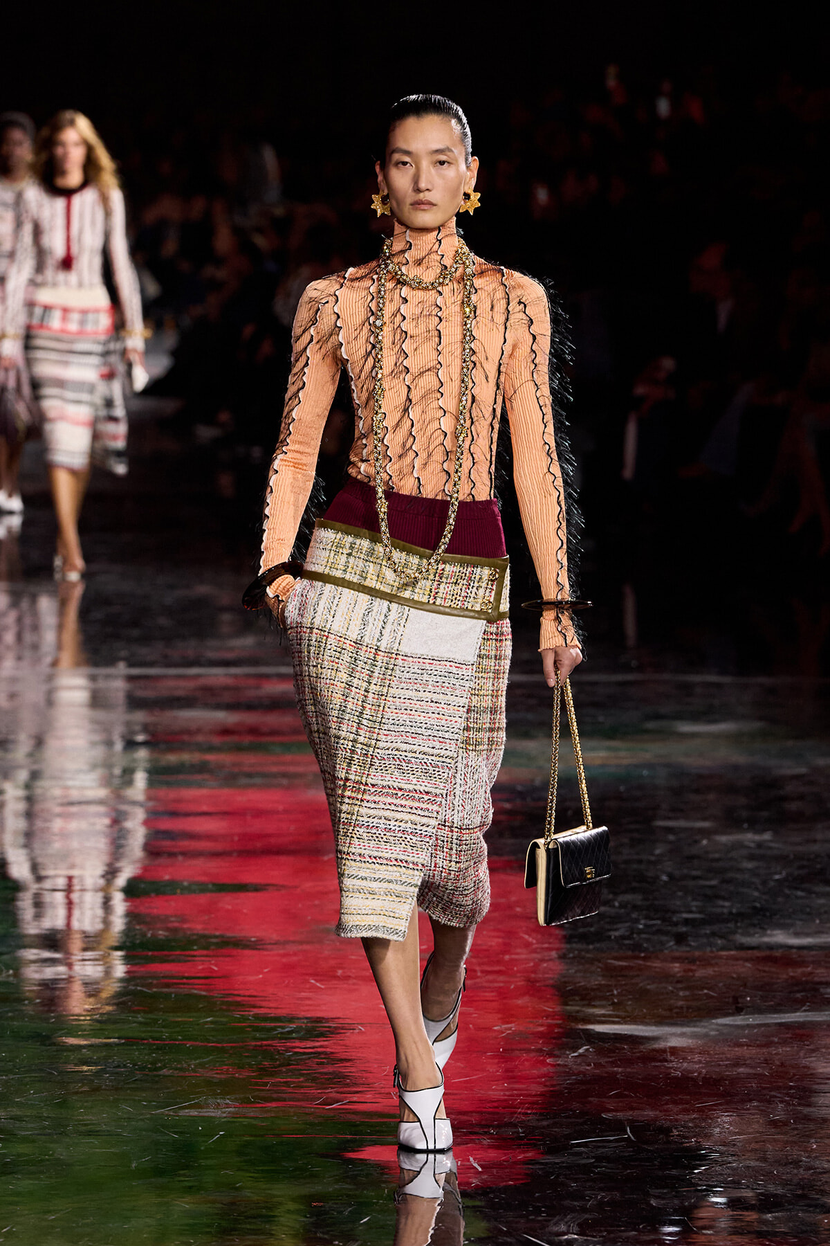 Model walking a runway in a peach sheer ribbed turtleneck with black trim, a plaid midi skirt, oversized gold jewelry, and a small black quilted handbag on a gold chain
