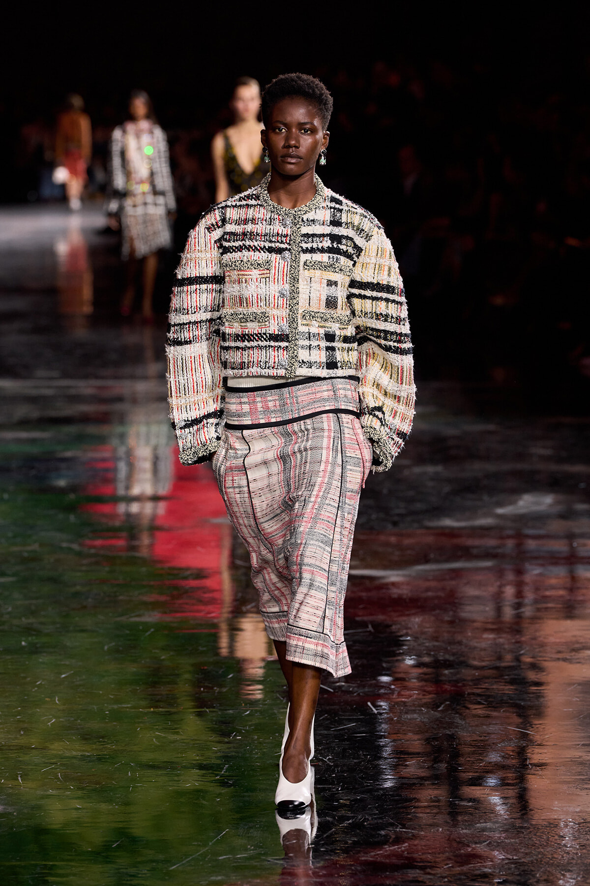 Model walks the runway in a textured tweed jacket and matching skirt with black, white, and subtle red/yellow threads; white heels, dark backstage setting.