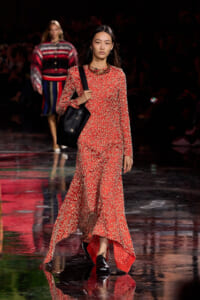 Runway model in a red floral long-sleeve maxi dress carrying a black quilted tote on a reflective catwalk.