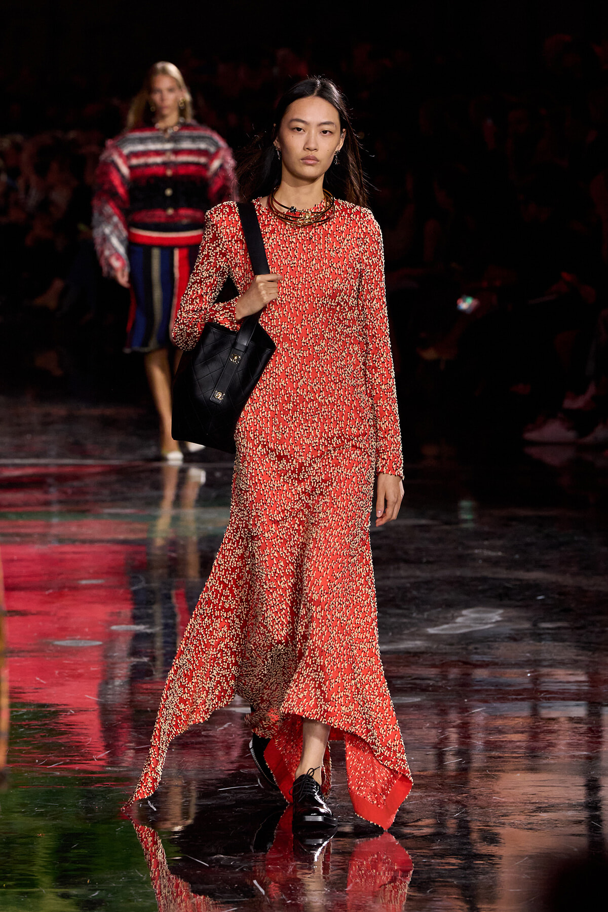 Runway model in a red floral long-sleeve maxi dress carrying a black quilted tote on a reflective catwalk.