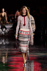 Model walking a fashion runway in a white knit top with red trim and a plaid midi skirt, holding a dark handbag.