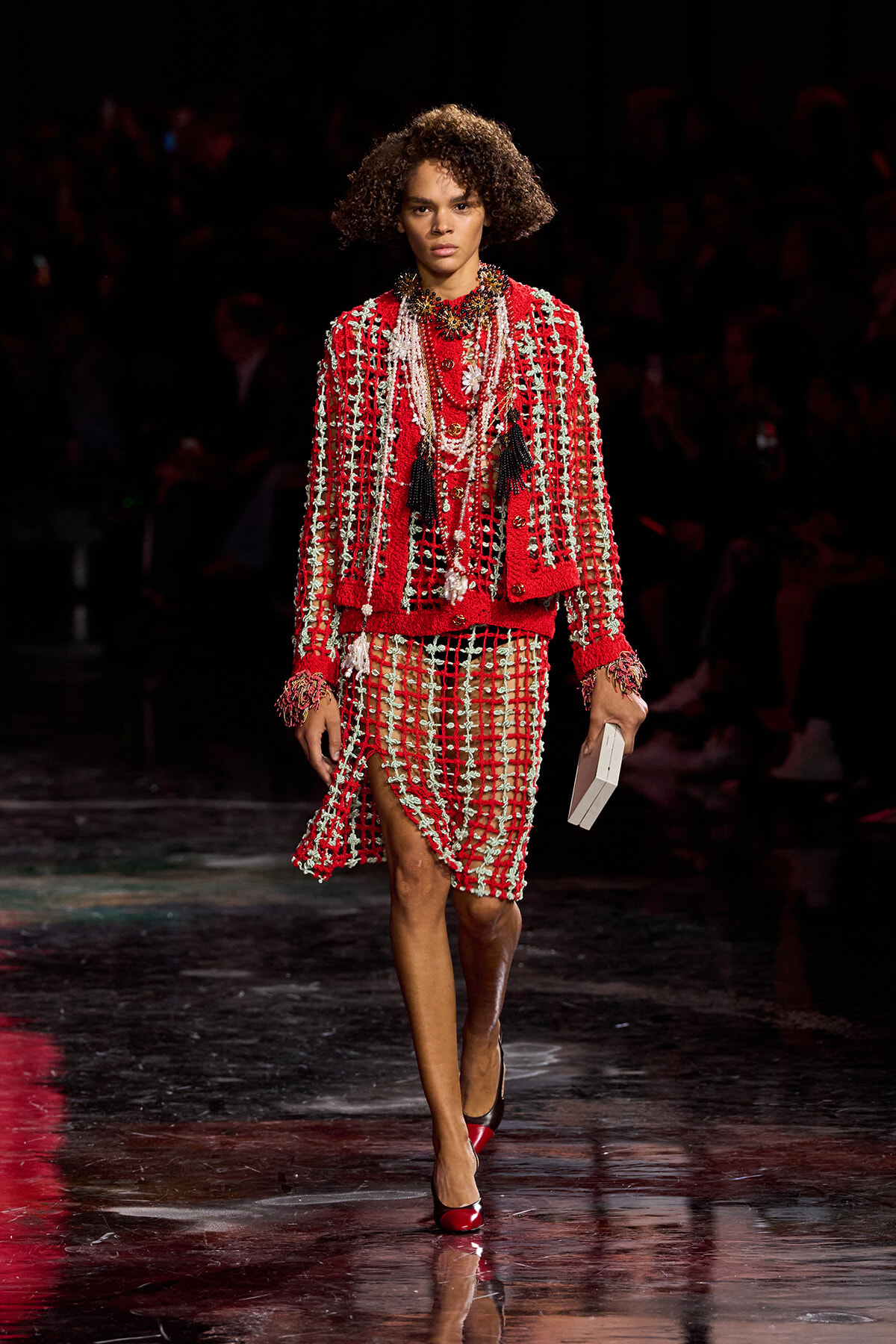 Model on a runway wearing a red beaded suit with white fringe and layered jewelry, holding a white clutch.