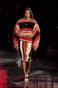 Model walking runway in a bold, multicolor, fringed coat with striped skirt and white-red heels, carrying a small white bag.
