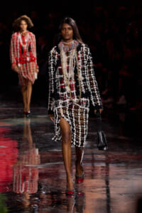 Runway model wearing a black knit coat with white fringe over a red plaid dress, layered pearl necklaces, and a black handbag.