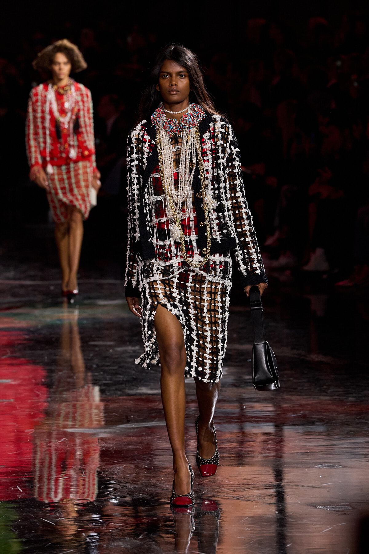 Runway model wearing a black knit coat with white fringe over a red plaid dress, layered pearl necklaces, and a black handbag.
