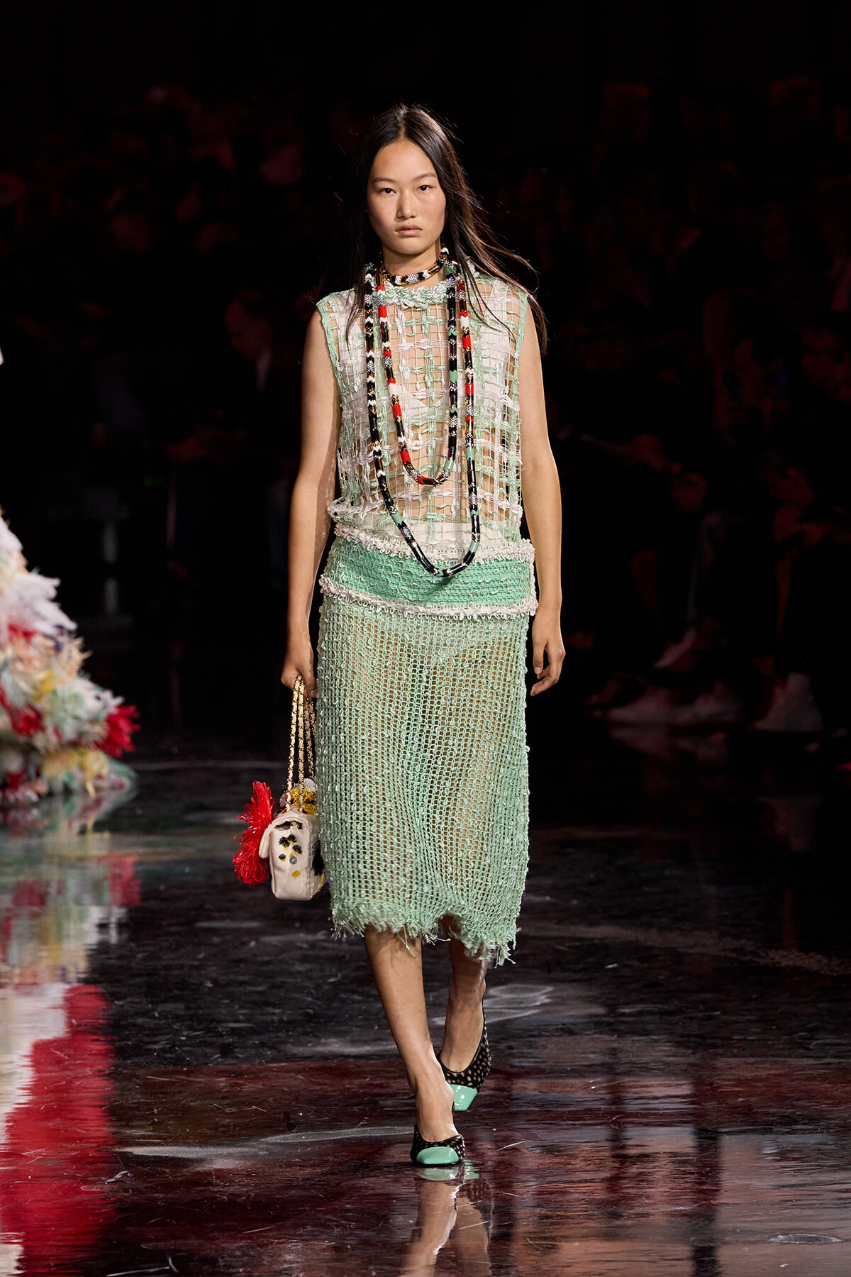 Model walking a runway in a mint-green, crocheted sleeveless dress with layered beaded necklaces and a small fringed handbag on her left.