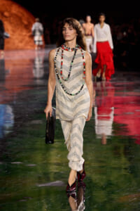 Model walks the runway in a sleeveless cream zigzag-knit jumpsuit, chunky beaded necklaces, and burgundy patent heels, carrying a black clutch.