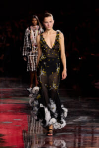 Model walks the runway in a sheer black gown with yellow floral embroidery and a feathered white-gray hem at the bottom.