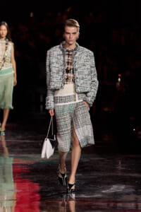 Model walks a runway in a cropped tweed jacket and matching skirt, carrying a white handbag and wearing black heels.