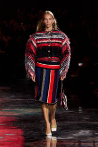 Model walking a fashion show runway in a multicolored, fringed knit sweater and matching striped skirt, carrying a striped handbag.